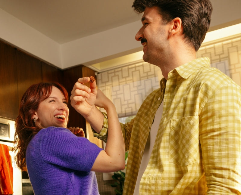 A joyful moment in a warmly lit kitchen where a man in a yellow plaid shirt playfully feeds a MadeGood Granola Bite to a woman in a purple sweater. She laughs with delight, leaning back slightly, as they share a fun, lighthearted interaction. The cozy setting and natural light add to the cheerful and relaxed atmosphere.