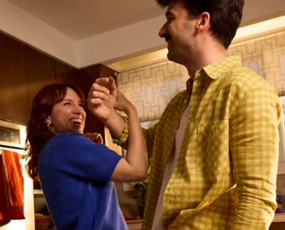 A joyful moment in a warmly lit kitchen where a man in a yellow plaid shirt playfully feeds a MadeGood Granola Bite to a woman in a purple sweater. She laughs with delight, leaning back slightly, as they share a fun, lighthearted interaction. The cozy setting and natural light add to the cheerful and relaxed atmosphere.