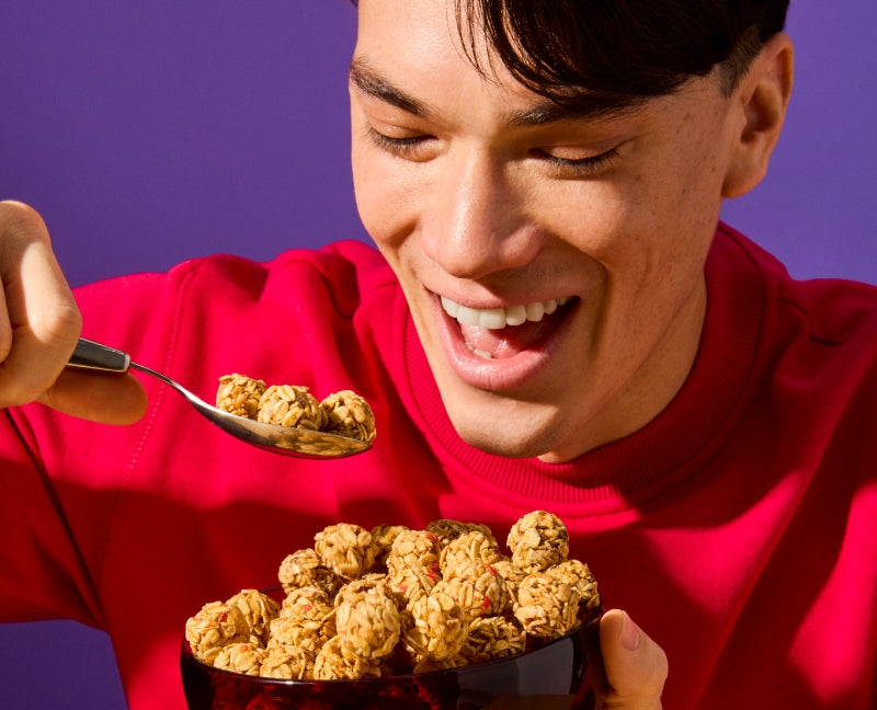 A cheerful young man in a red sweatshirt is holding a bowl filled with MadeGood Granola Bites. He is smiling excitedly as he scoops up a spoonful, ready to take a bite. One snack bite appears to be mid-air, adding a playful touch. The vibrant purple background contrasts with his red outfit, making the scene feel lively and energetic.