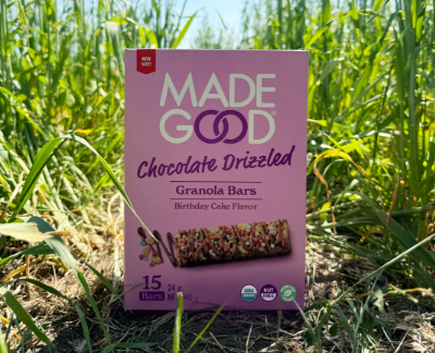 Box of "Made Good" chocolate drizzled granola bars with birthday cake flavor in a lush field. Vibrant greenery surrounds the purple packaging.