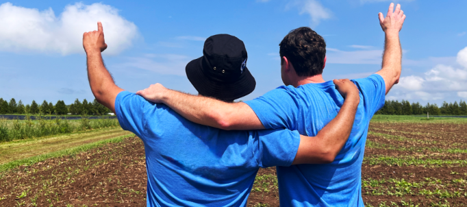 Two people in blue shirts stand with arms around each other, facing a field. They raise their arms, conveying friendship and celebration under a blue sky.