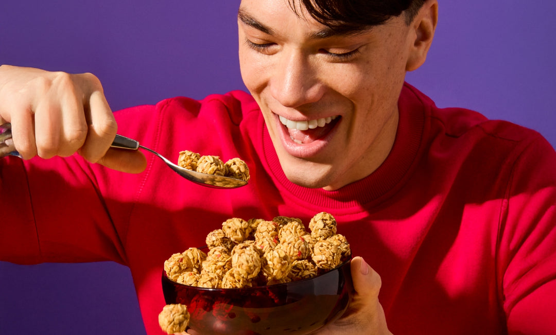 A cheerful young man in a red sweatshirt is holding a bowl filled with MadeGood Granola Bites. He is smiling excitedly as he scoops up a spoonful, ready to take a bite. One snack bite appears to be mid-air, adding a playful touch. The vibrant purple background contrasts with his red outfit, making the scene feel lively and energetic.