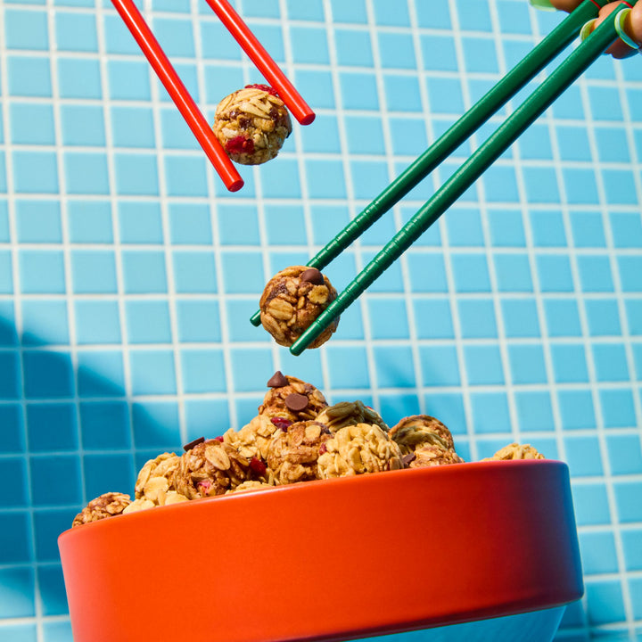 A playful and colorful scene featuring a bright orange bowl filled with MadeGood Granola Bites against a blue tiled background. Two sets of chopsticks, one green and one red, are picking up snack bites, with one bite appearing to be mid-air. The contrasting colors and dynamic composition create a fun, energetic feel.