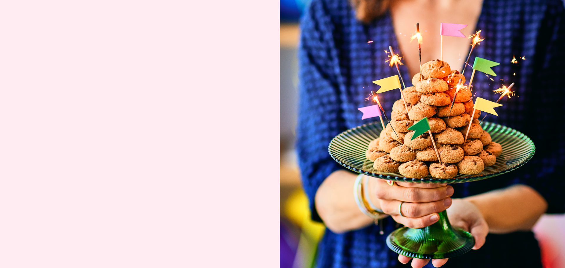 A person in a blue outfit holds a green glass cake stand with a festive cookie tower, decorated with colorful flags and sparklers for a celebratory touch.