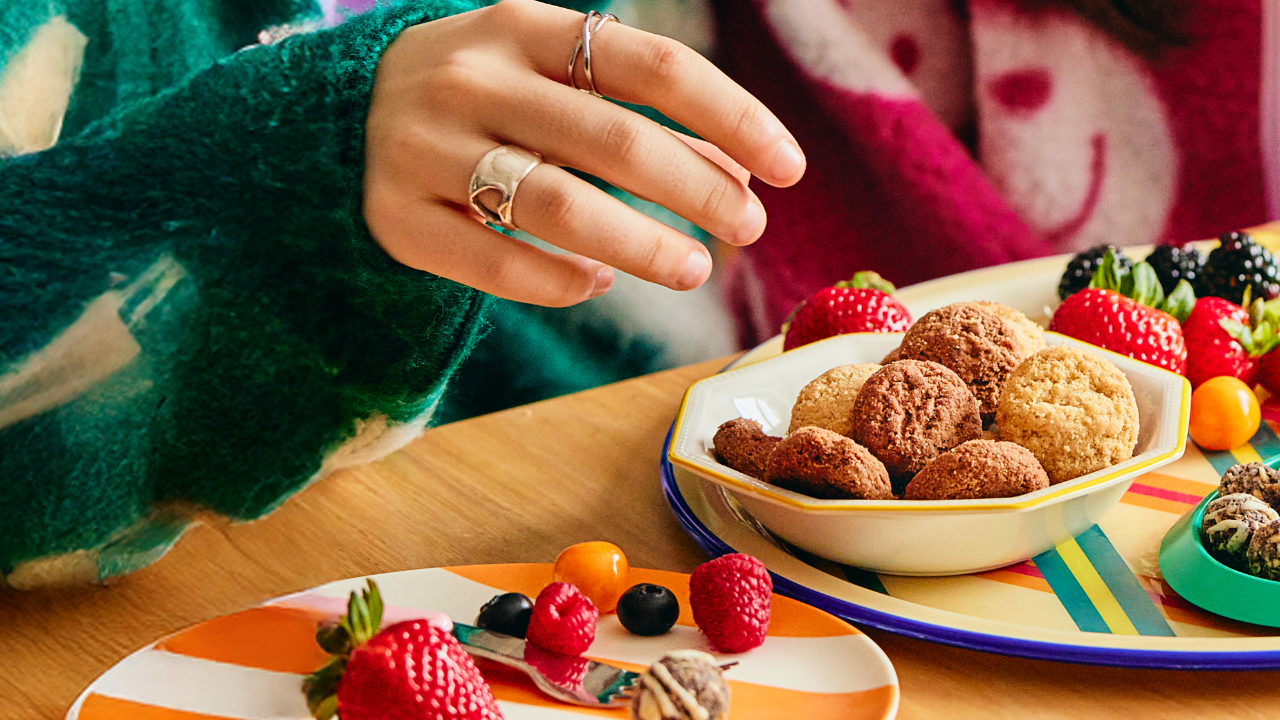 A hand reaches for cookies in a bowl surrounded by colorful fruit like strawberries and berries. The scene conveys a cozy and inviting atmosphere.