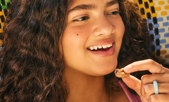 Smiling young woman with curly hair holds a small cookie near her mouth. She is joyful and sits in front of a colorful mosaic background.