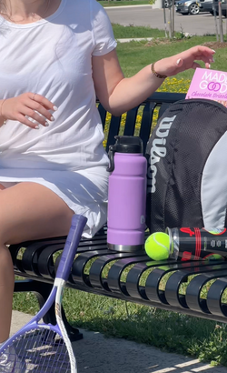 A person in a white dress sitting on a park bench, holding a pink box of MadeGood Chocolate Drizzled Bars. Beside them is a purple water bottle, a black and white Wilson backpack, a canister of tennis balls, and a purple tennis racket resting against the bench.