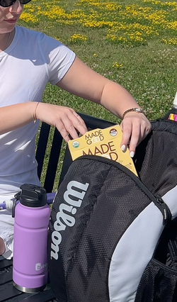 A person wearing a white shirt placing a yellow box of MadeGood snacks into a black and white Wilson backpack, with a purple water bottle beside them on a bench and a field of yellow flowers in the background.