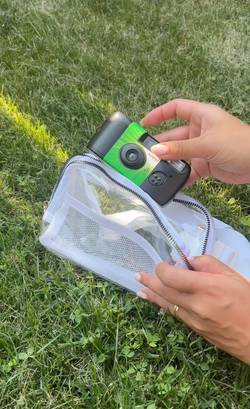 A person placing a green and black disposable Fujifilm camera into a clear, zippered pouch while sitting on lush green grass.