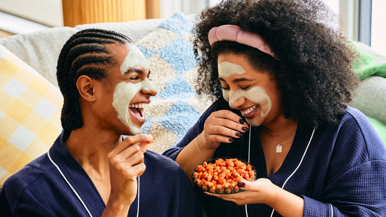 Two friends wearing matching navy blue robes and green facial masks are sitting on a couch, laughing and snacking. One person holds a small bowl filled with MadeGood Star Puffed Crackers, while the other reaches for a piece. They are relaxed, enjoying a cozy self-care moment with drinks on the table.