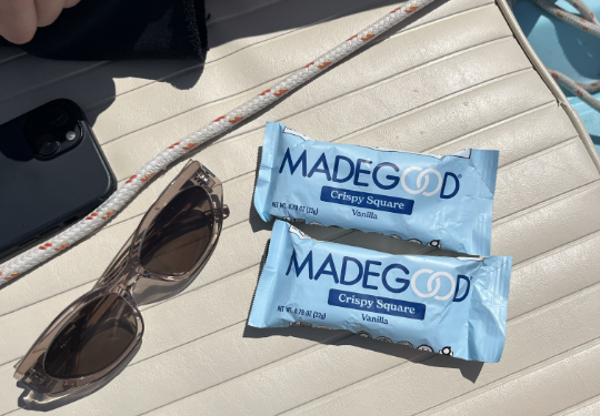 Two MadeGood crispy squares in blue packaging on a sunlit padded surface, accompanied by sunglasses and a smartphone. Bright and relaxed vibe.