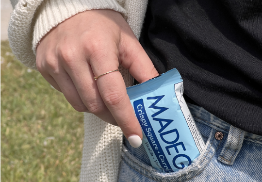 A hand with white nail polish and a ring slips a blue MADE GOOD snack bar into a pocket of faded jeans. The background is grassy and sunlit.