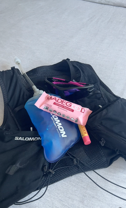 A pink MadeGood Strawberry Crispy Square bar rests on top of a blue Salomon hydration flask, which is placed in a black running vest. Nearby, a pair of purple-lensed sunglasses and a tube of Burt's Bees lip balm are visible, suggesting essentials for an active, outdoor adventure.