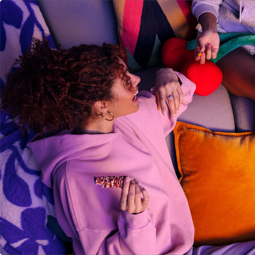 A woman in a pink hoodie lounges on a cozy couch, holding a colorful granola bar with a drizzle topping. She is engaged in conversation with another person, who is offering her a snack. The setting is warm and relaxed, with vibrant pillows and a plush cherry-shaped cushion adding to the cozy atmosphere.