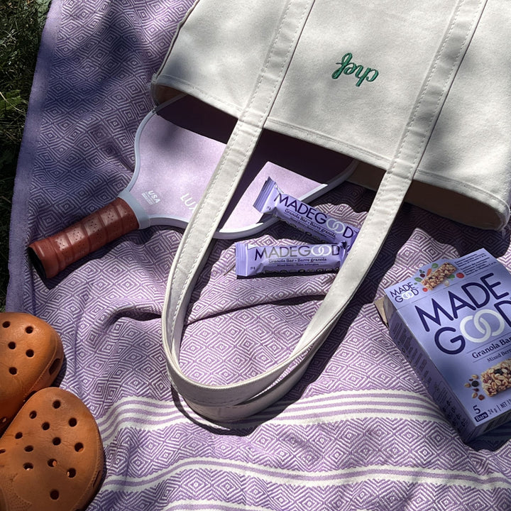 A neatly arranged outdoor picnic scene features a lavender-patterned blanket spread on the grass. A white canvas tote bag with green embroidery rests on top, partially covering a pastel purple pickleball paddle. Two MadeGood granola bars in lavender packaging lay beside the paddle, along with a box of MadeGood granola bars. A pair of tan perforated clogs sits nearby, completing the relaxed and sunlit setup.