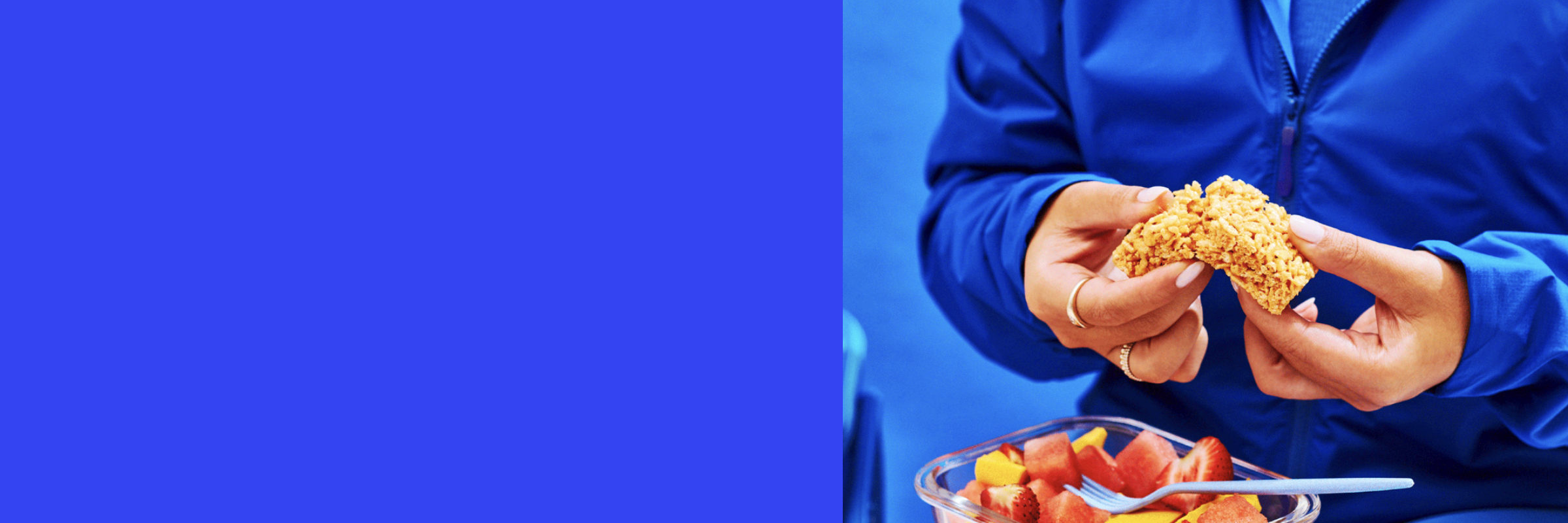 A person in a blue tracksuit breaks apart a crispy square treat while holding a glass container filled with fresh fruit, including watermelon, mango, and strawberries.