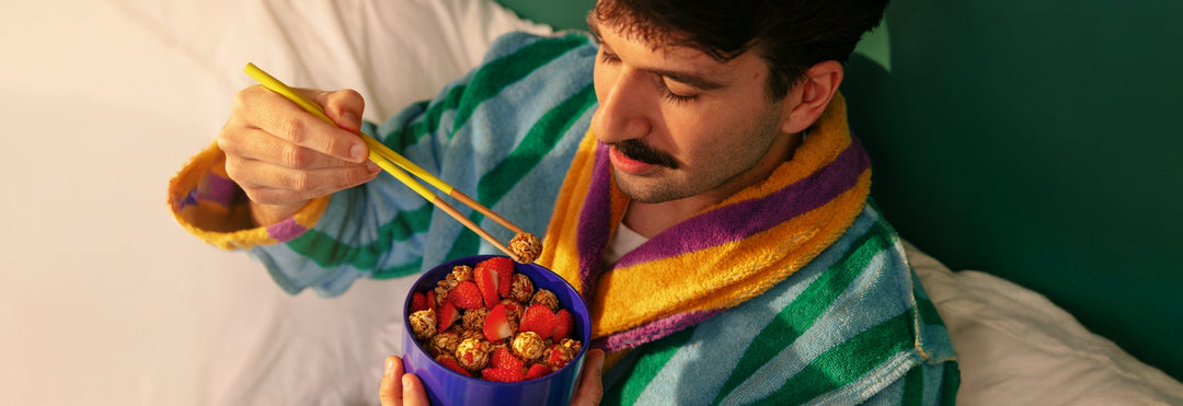 Man in a colorful robe uses yellow chopsticks to eat granola and strawberries from a blue bowl. The setting is cozy, conveying a relaxed morning vibe.