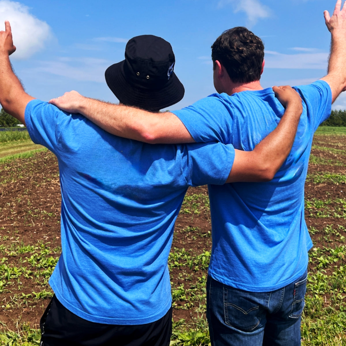 Two people in blue shirts stand with arms around each other, facing a field. They raise their arms, conveying friendship and celebration under a blue sky.