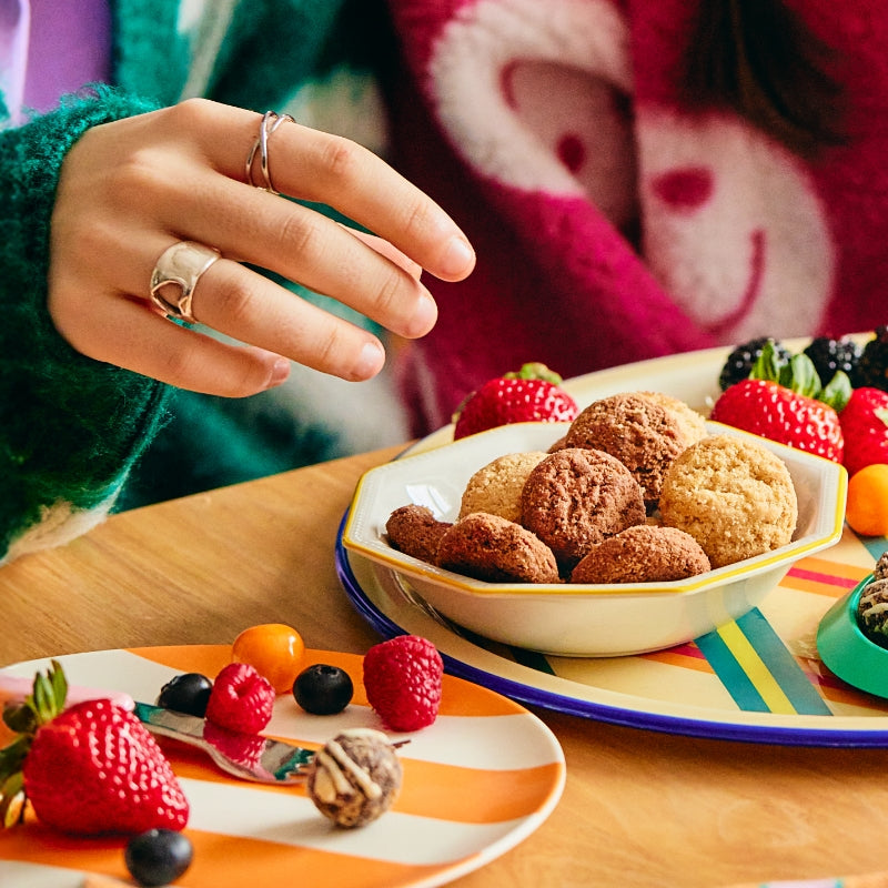 A hand reaches for a cookie placed in a bowl amongst other fruits on colorful striped plates.