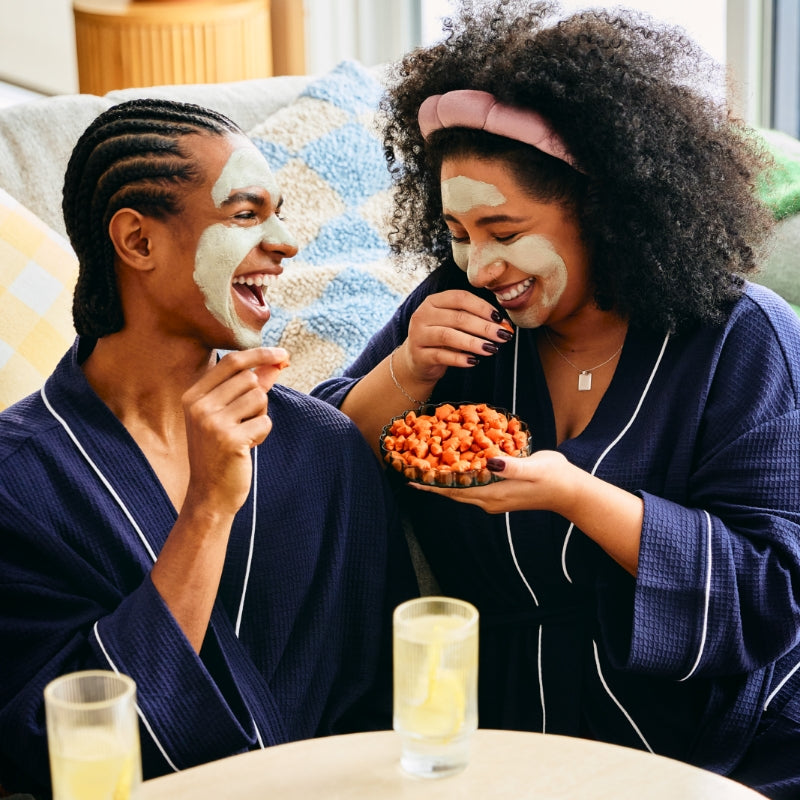 Two friends wearing matching navy blue robes and green facial masks are sitting on a couch, laughing and snacking. One person holds a small bowl filled with MadeGood Star Puffed Crackers, while the other reaches for a piece. They are relaxed, enjoying a cozy self-care moment with drinks on the table.