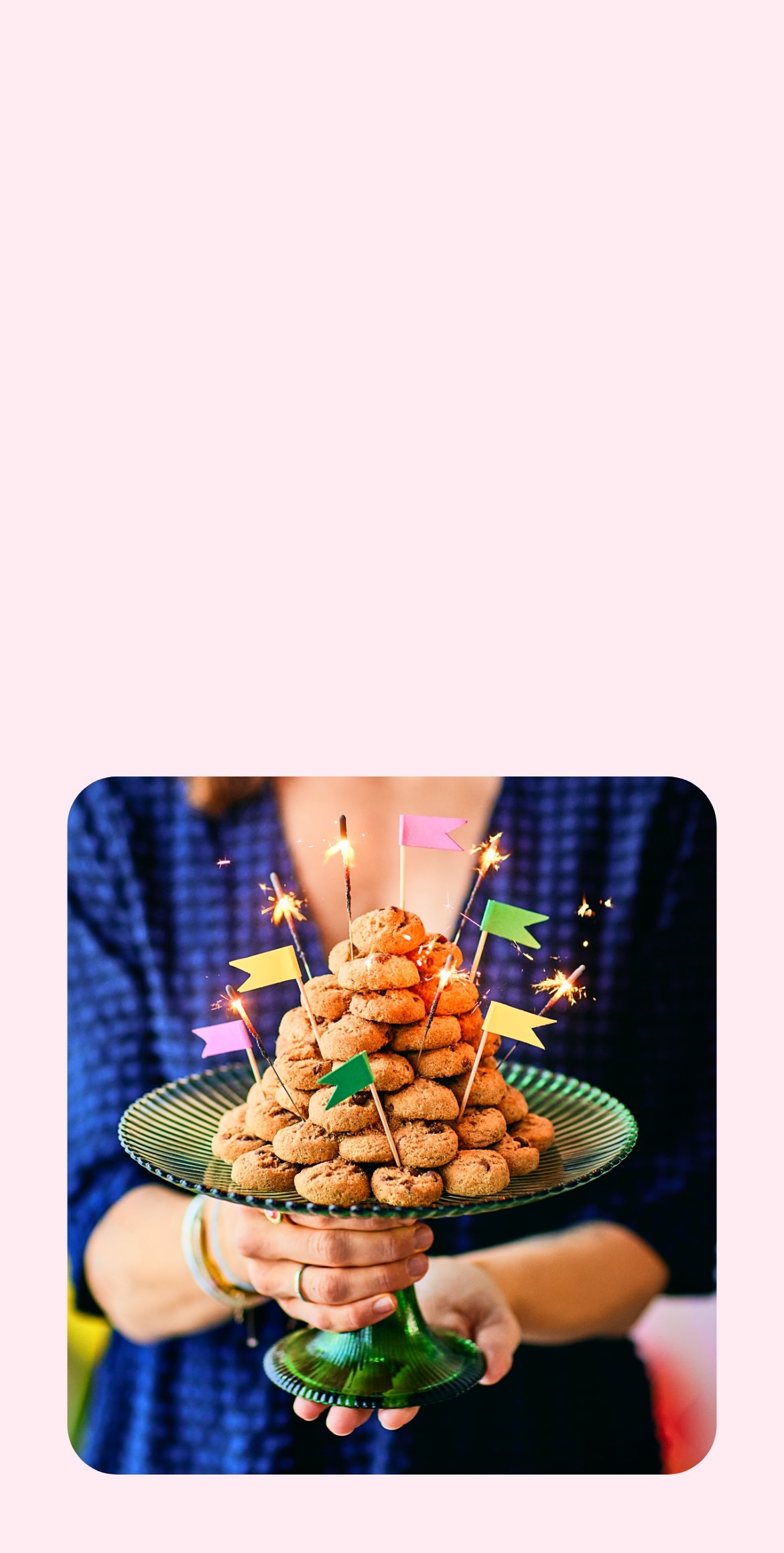 A person in a blue outfit holds a green glass cake stand with a festive cookie tower, decorated with colorful flags and sparklers for a celebratory touch.