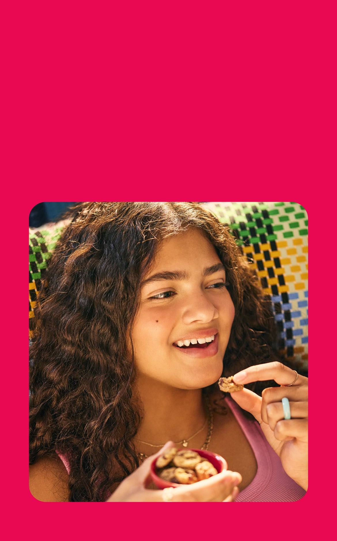 A young woman with curly brown hair reclines on a colorful woven chair, smiling as she enjoys a bite-sized soft-baked cookie. She holds a small red bowl filled with more cookies in one hand, while the other lifts a piece to her mouth. She wears a pink tank top, layered gold necklaces, and a turquoise ring, basking in the warm sunlight.