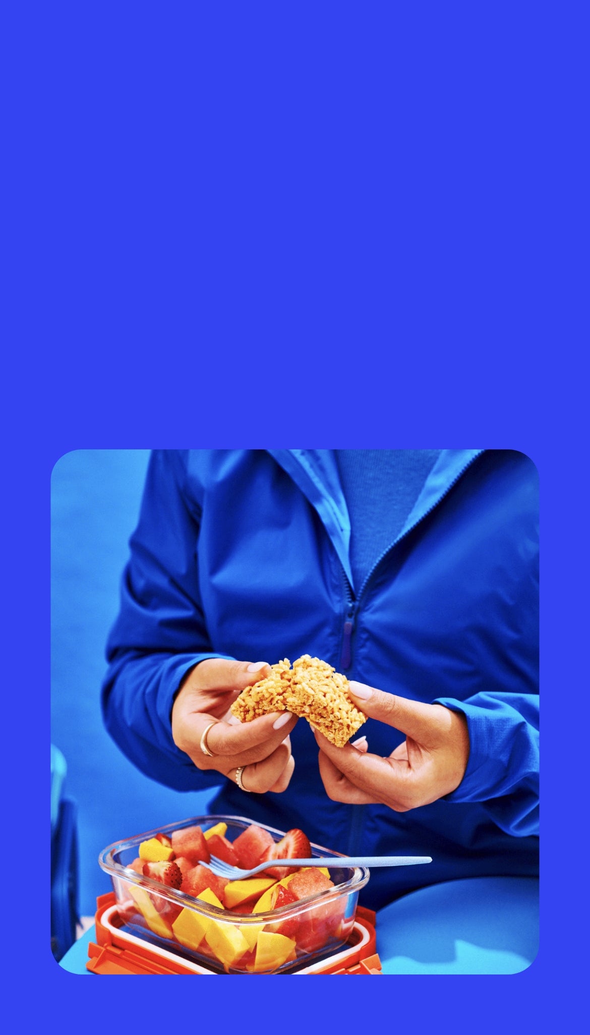 A person in a blue tracksuit breaks apart a crispy square treat while holding a glass container filled with fresh fruit, including watermelon, mango, and strawberries.