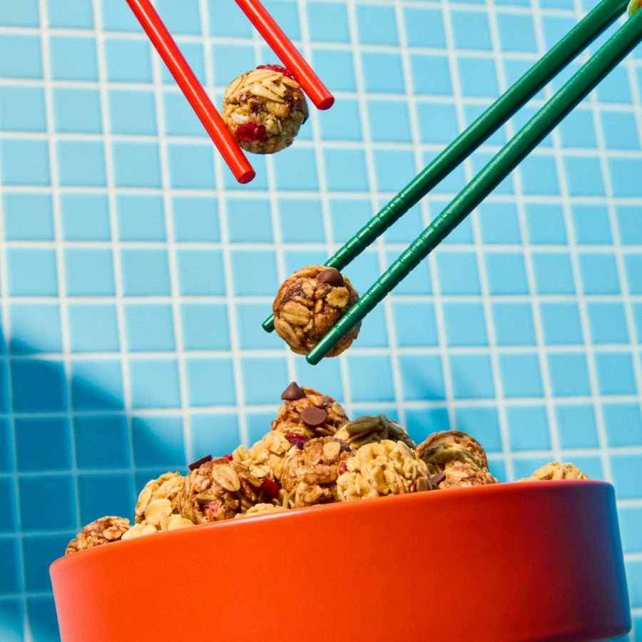 A playful and colorful scene featuring a bright orange bowl filled with MadeGood Granola Bites against a blue tiled background. Two sets of chopsticks, one green and one red, are picking up snack bites, with one bite appearing to be mid-air. The contrasting colors and dynamic composition create a fun, energetic feel.