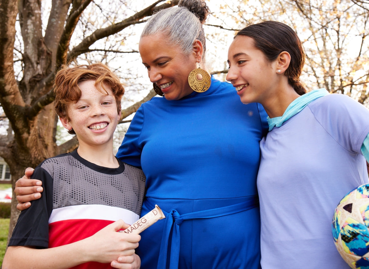 A woman in a blue dress embraces a smiling boy and girl holding a soccer ball. They stand outdoors under a tree, exuding warmth and happiness.