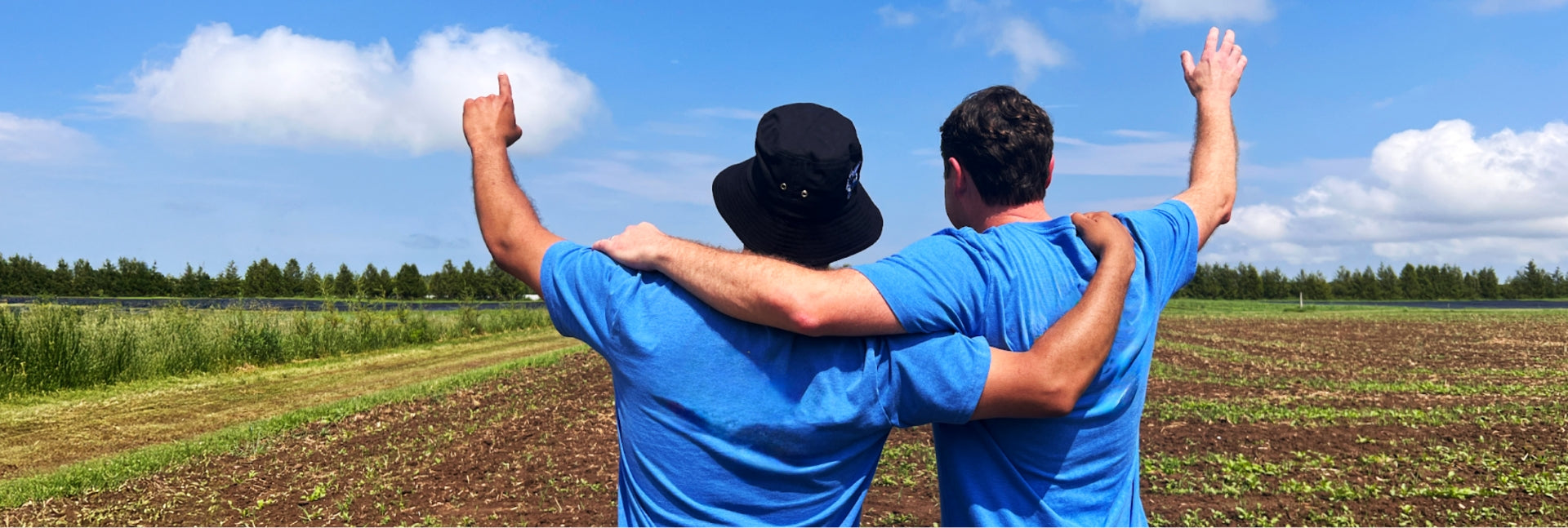 Two men stand in the middle of a field and clear blue skies with am arm on each other's shoulders with the other arm reaching upwards to the sky.