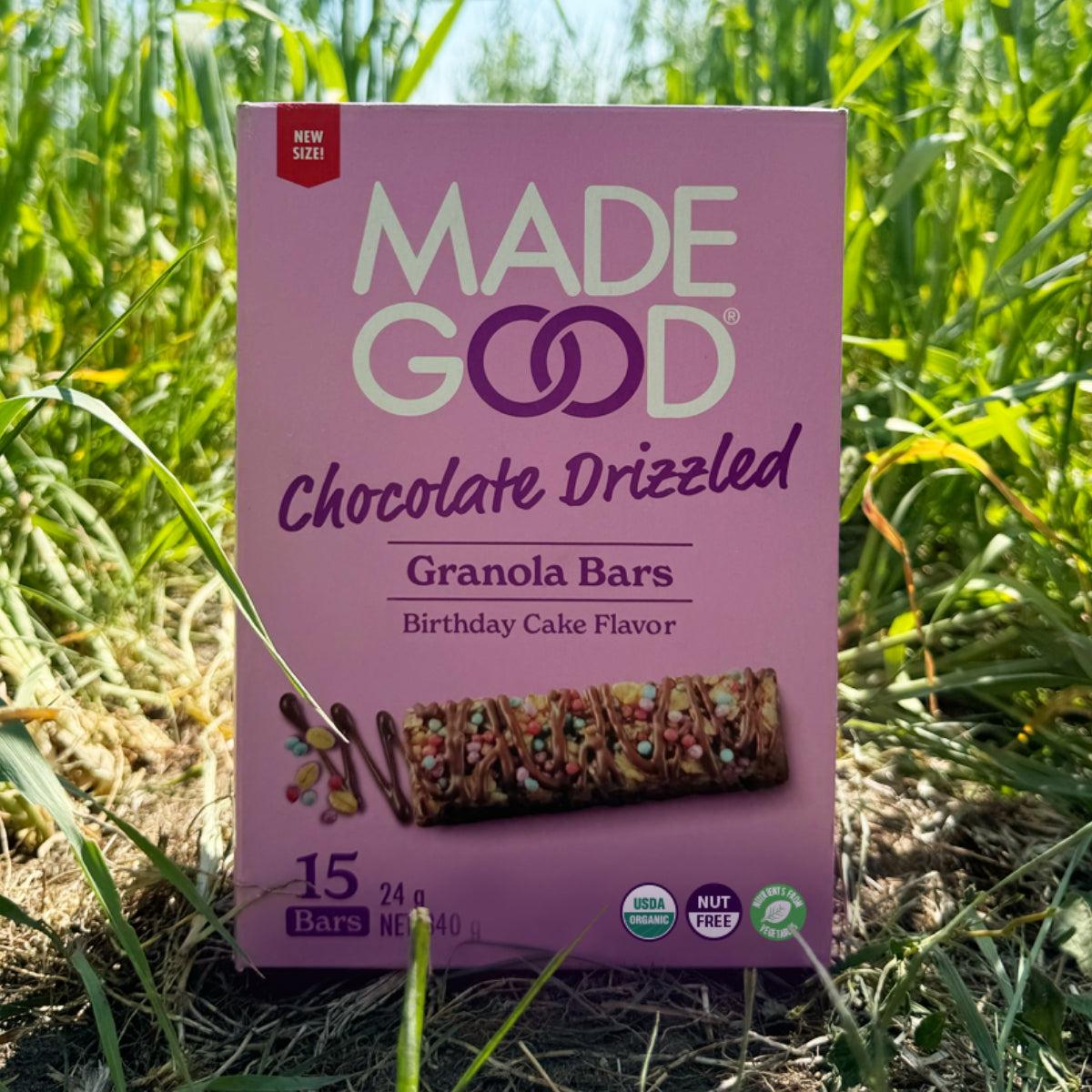 Box of Made Good Chocolate Drizzled Granola Bars, Birthday Cake flavor, in a grassy field. The package is purple with bright, colorful accents.