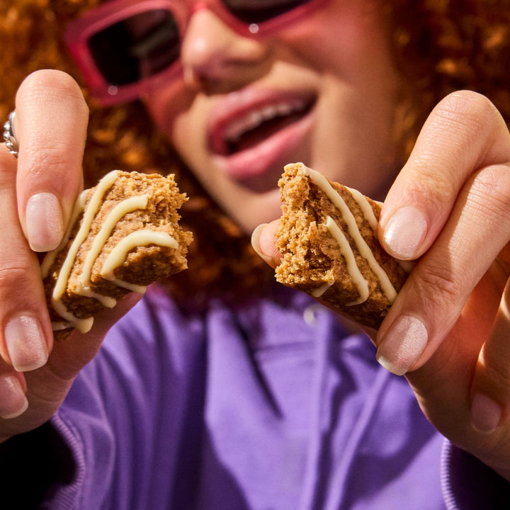 A young woman with curly red hair and pink sunglasses smiles as she holds and breaks apart a Mornings soft baked breakfast oat bar. She is wearing a purple sweater, and the bar’s soft, crumbly texture is visible, with icing drizzled in a zigzag pattern.