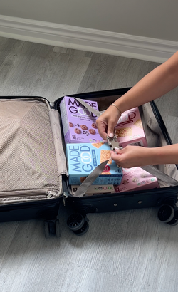 A person buckling the straps of a suitcase filled with MadeGood snack boxes, including Mini Biscuits, Crispy Squares, and Granola Minis, neatly packed on a light wood floor.