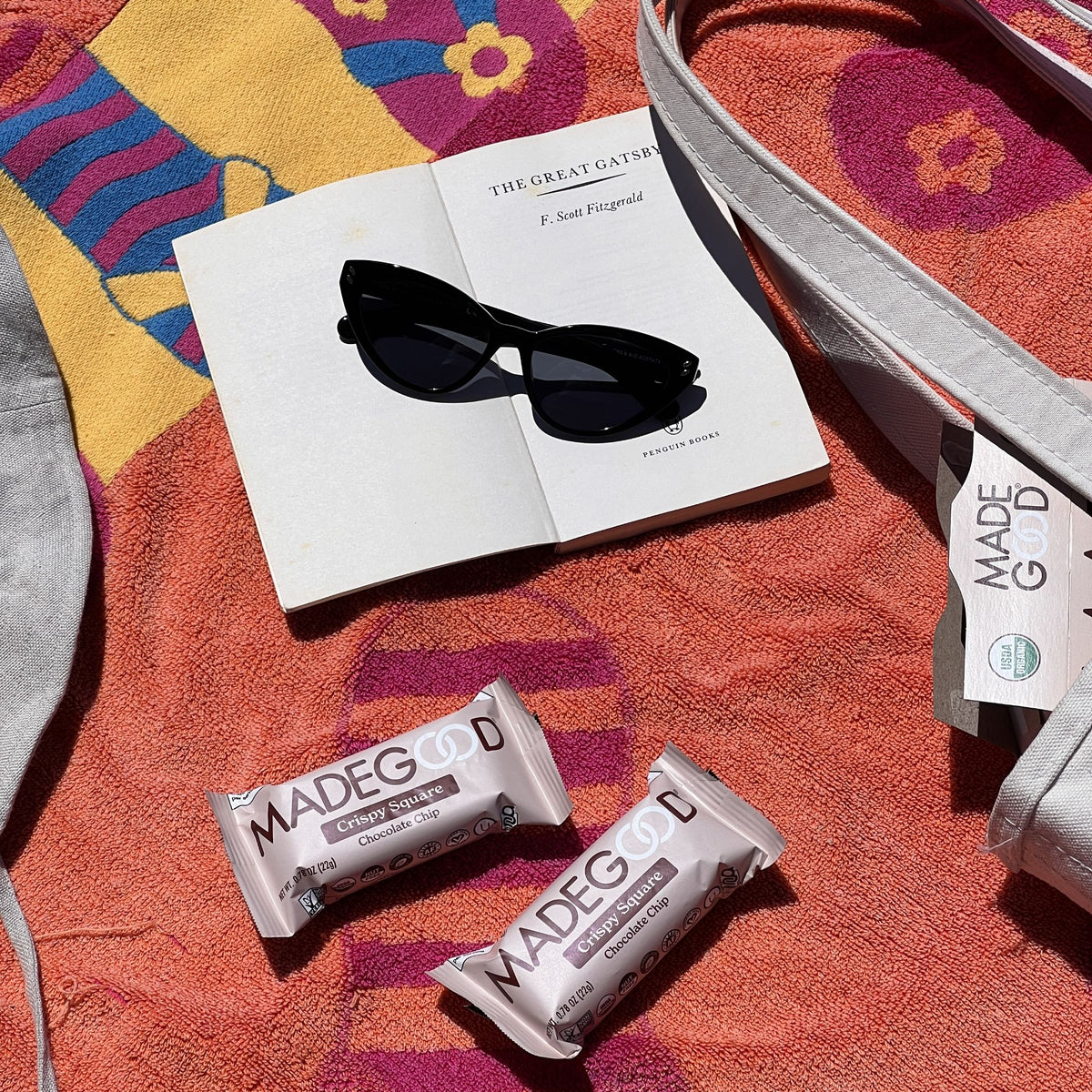 A sunny beachside setup featuring an open book, black cat-eye sunglasses, and two MadeGood Chocolate Chip Crispy Squares resting on a colorful towel. A white tote bag with a MadeGood tag peeks into the frame.