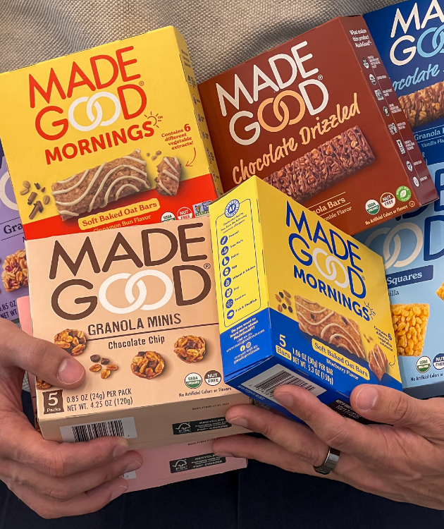 A person holds various "Made Good" snack boxes, including granola minis and oat bars. The bright packaging suggests a healthy, vibrant snack option.