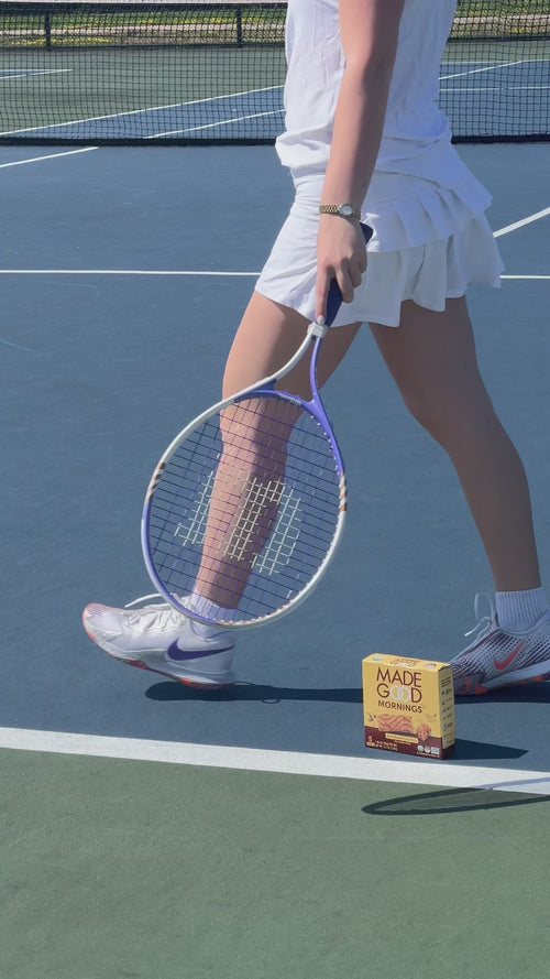 A woman walks across a tennis court with a box of MadeGood Mornings bars on court. Transitions to her seated on a park bench eating the mornings bar.